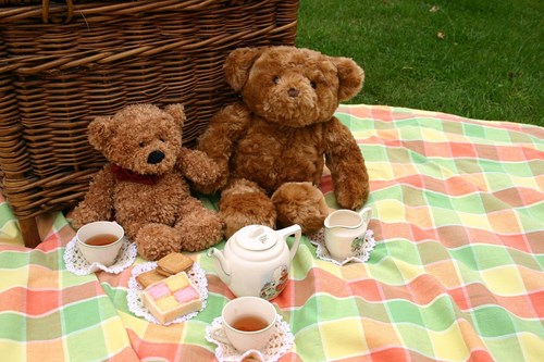 Two teddy bears sat in front of a picnic basket on a blanket with tea set