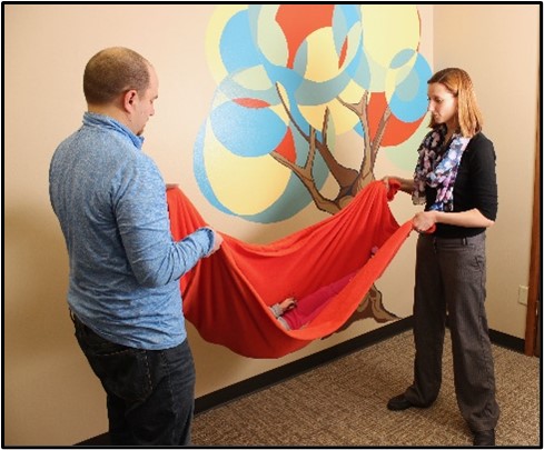 Picture of two people holding another person in an orange sling with a picture of a tree in the background